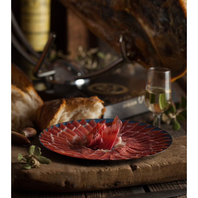  A beautifully plated serving of Cinco Jotas 5J Iberico Jamon, with bread and a glass of wine in the background.